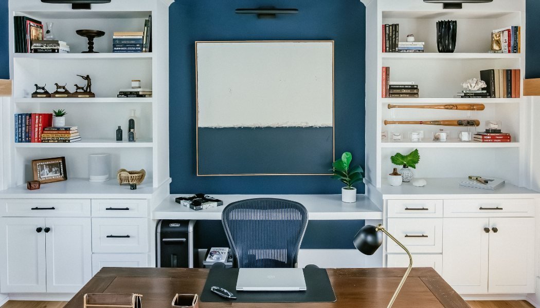 Home office with navy accent wall, white built-in shelving and a walnut desk