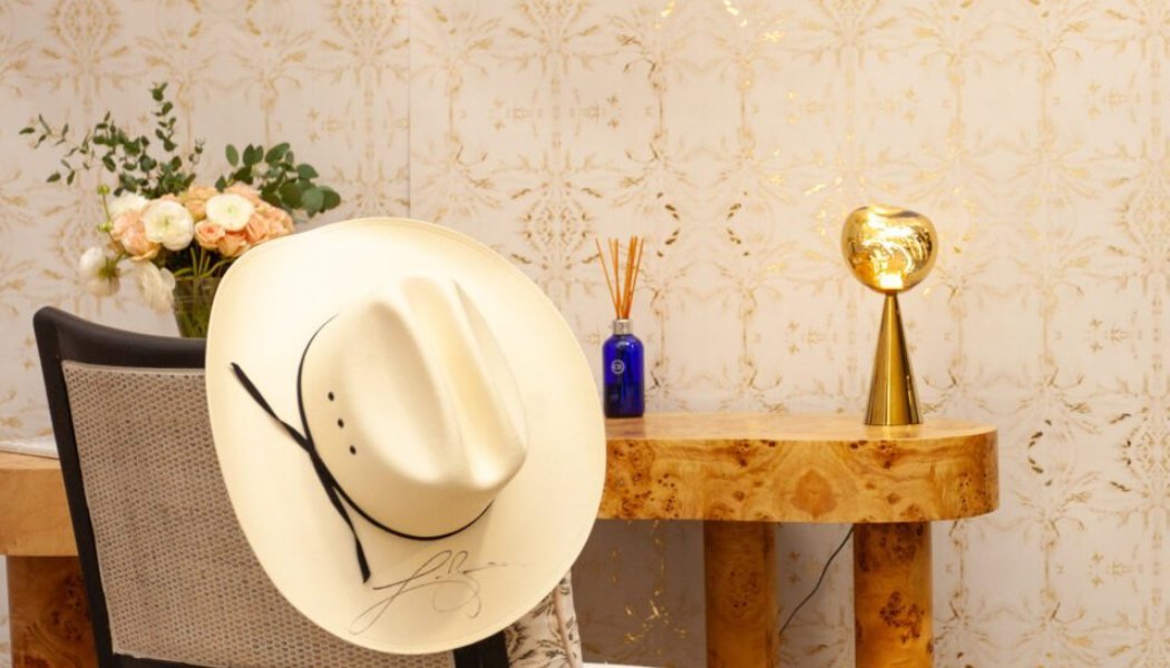 Signed cream cowboy hat beside a burl-wood console and gold lamp against gold damask wallpaper