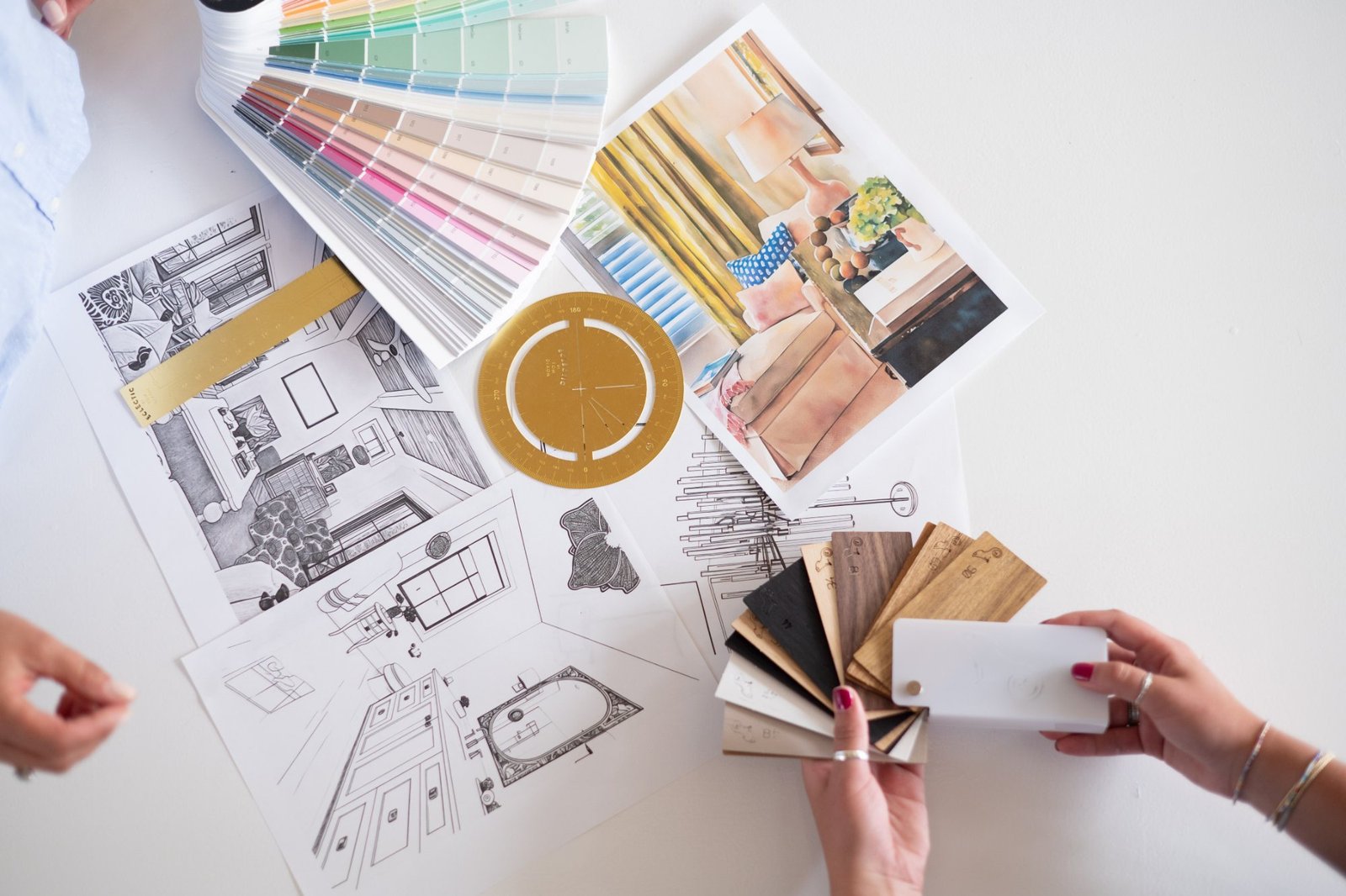 Two designers’ hands fanning paint chips and wood samples over floor plans, sketches, and a watercolor rendering
