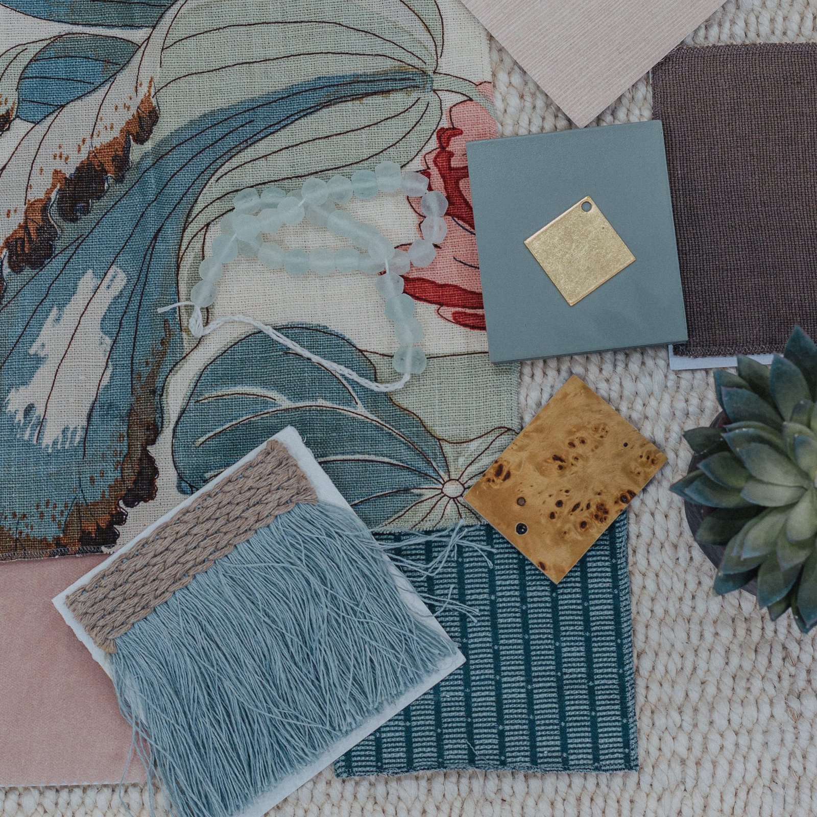 A flat lay of fabric and finish samples — a botanical print, sea-glass beads, sage and burl wood chips, blue-green weaves, and fringe trim on a woven jute rug