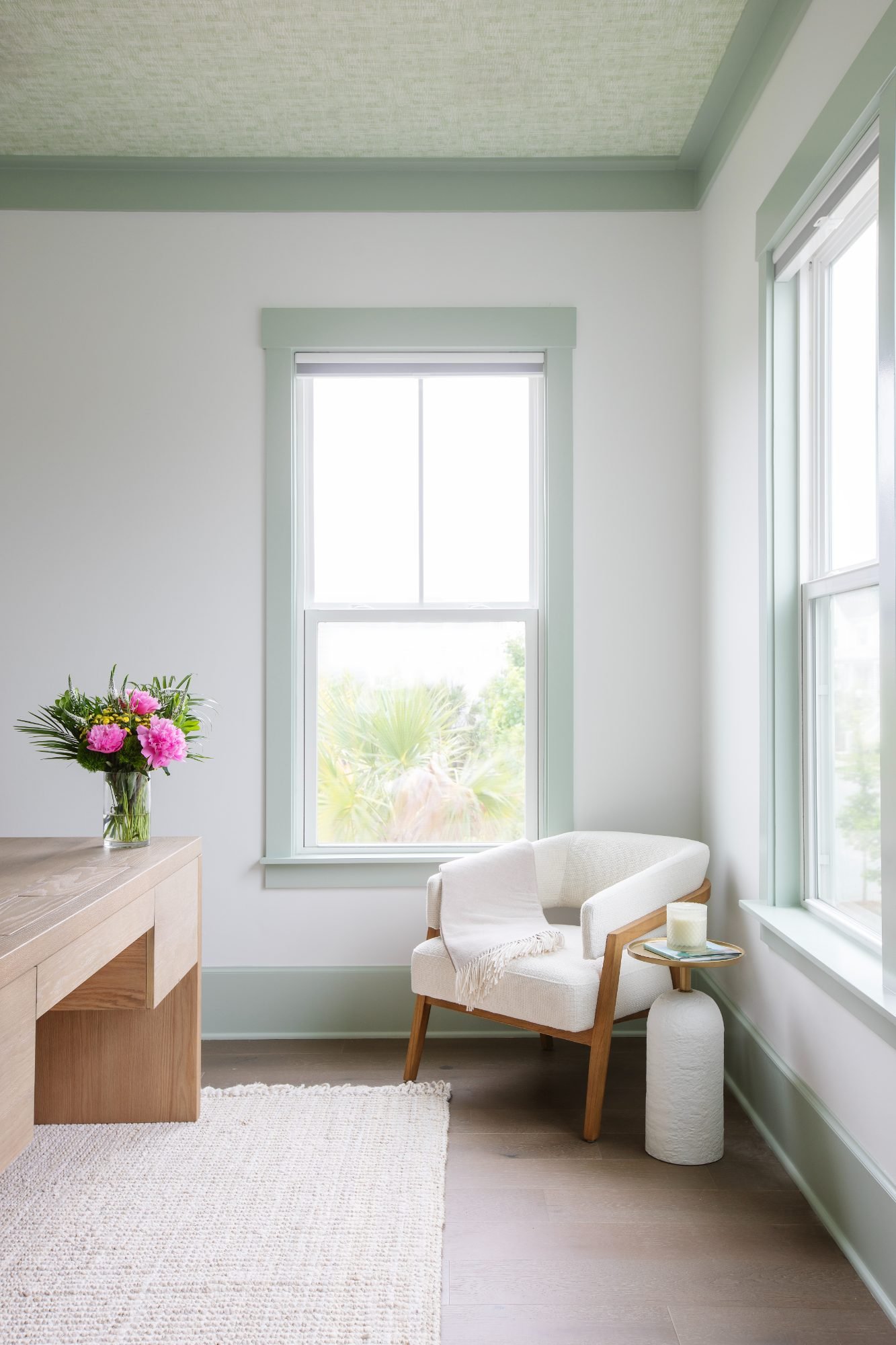 A quiet office corner with sage-green trim, a white bouclé armchair, and peonies on a white-oak desk