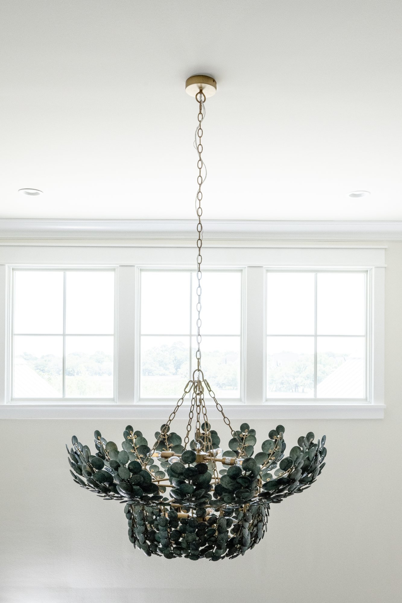 A tiered chandelier of dark green capiz discs on an aged brass frame, hung before three white-trimmed windows