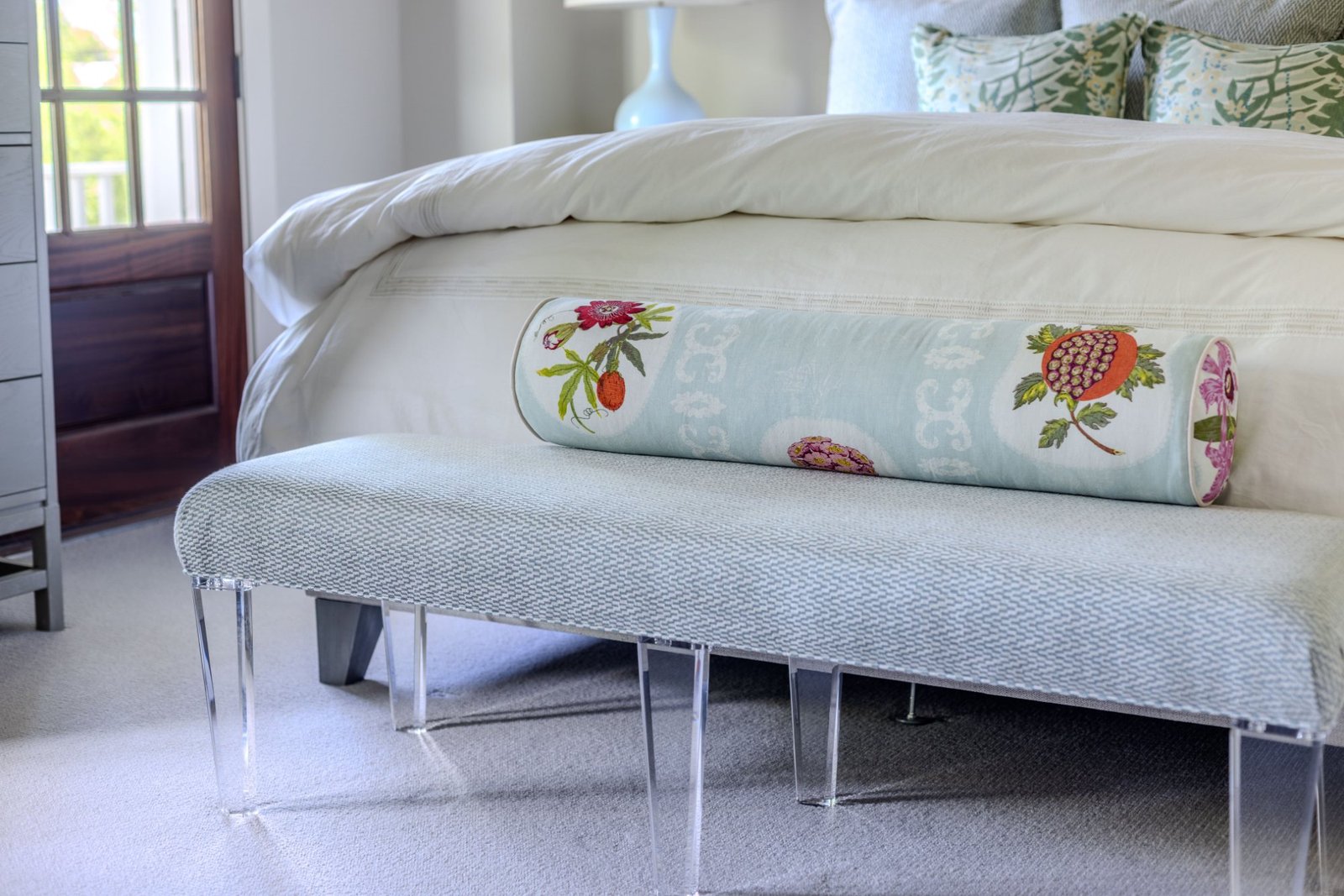 Bolster resting on a woven bench with acrylic legs at the foot of the bed