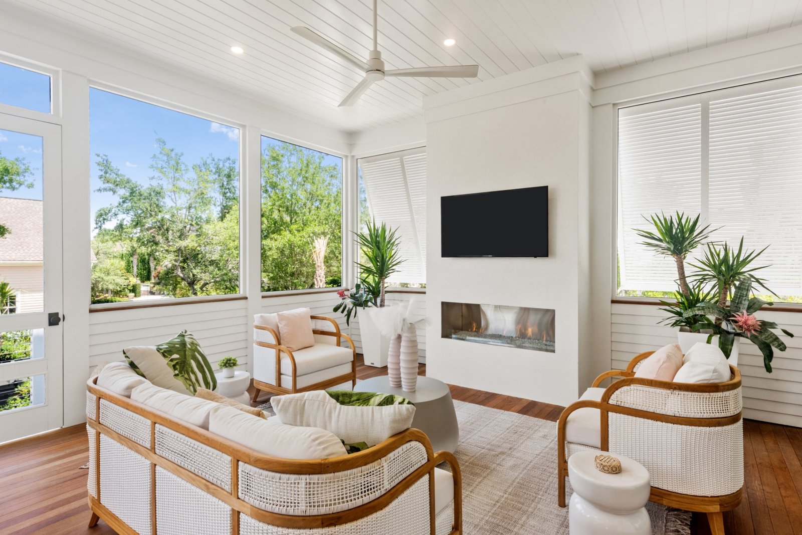 A screened porch with white shiplap walls, rattan seating with cream cushions, a linear fireplace, and marsh-side greenery through the windows