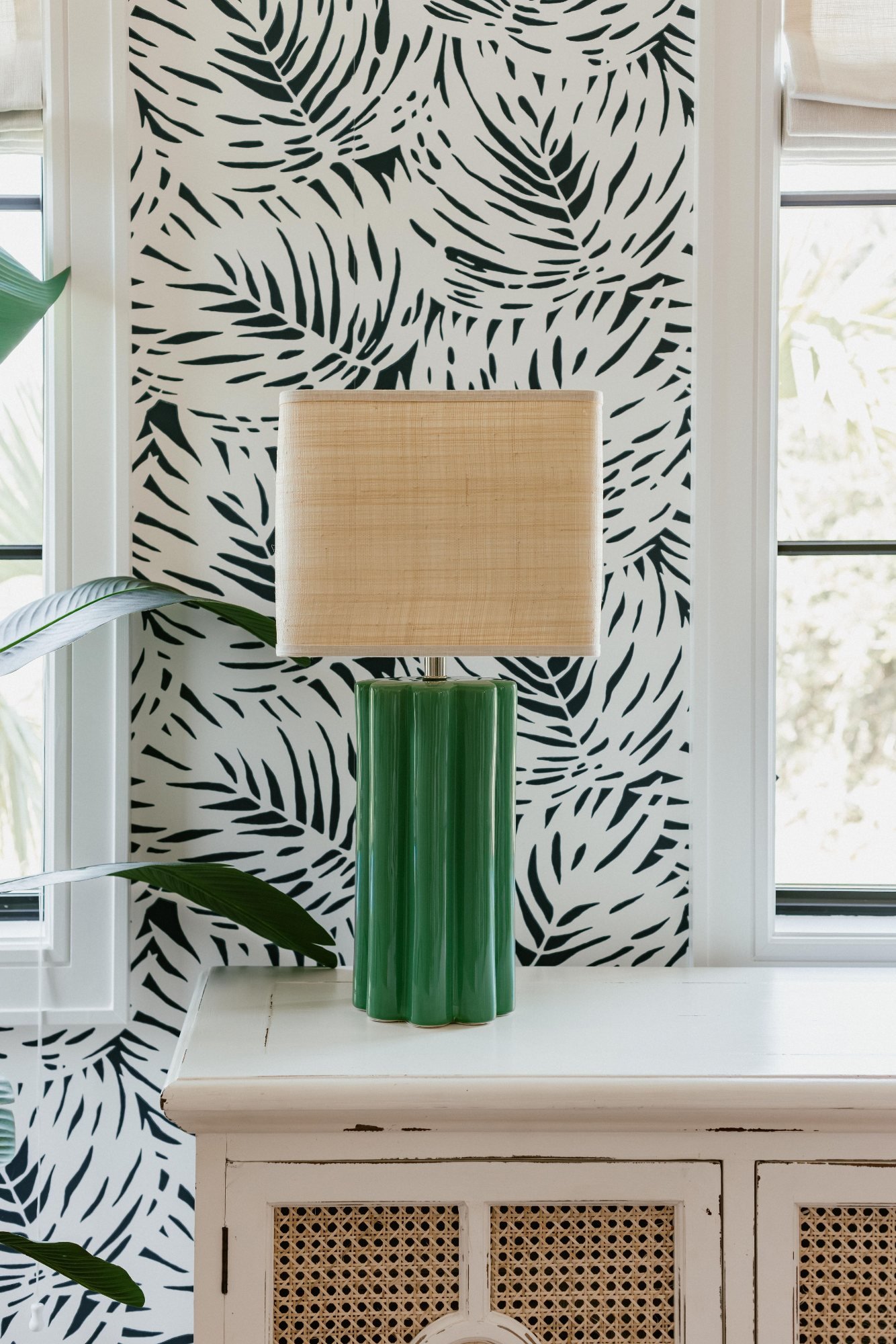 Green fluted ceramic lamp with raffia shade against palm wallpaper