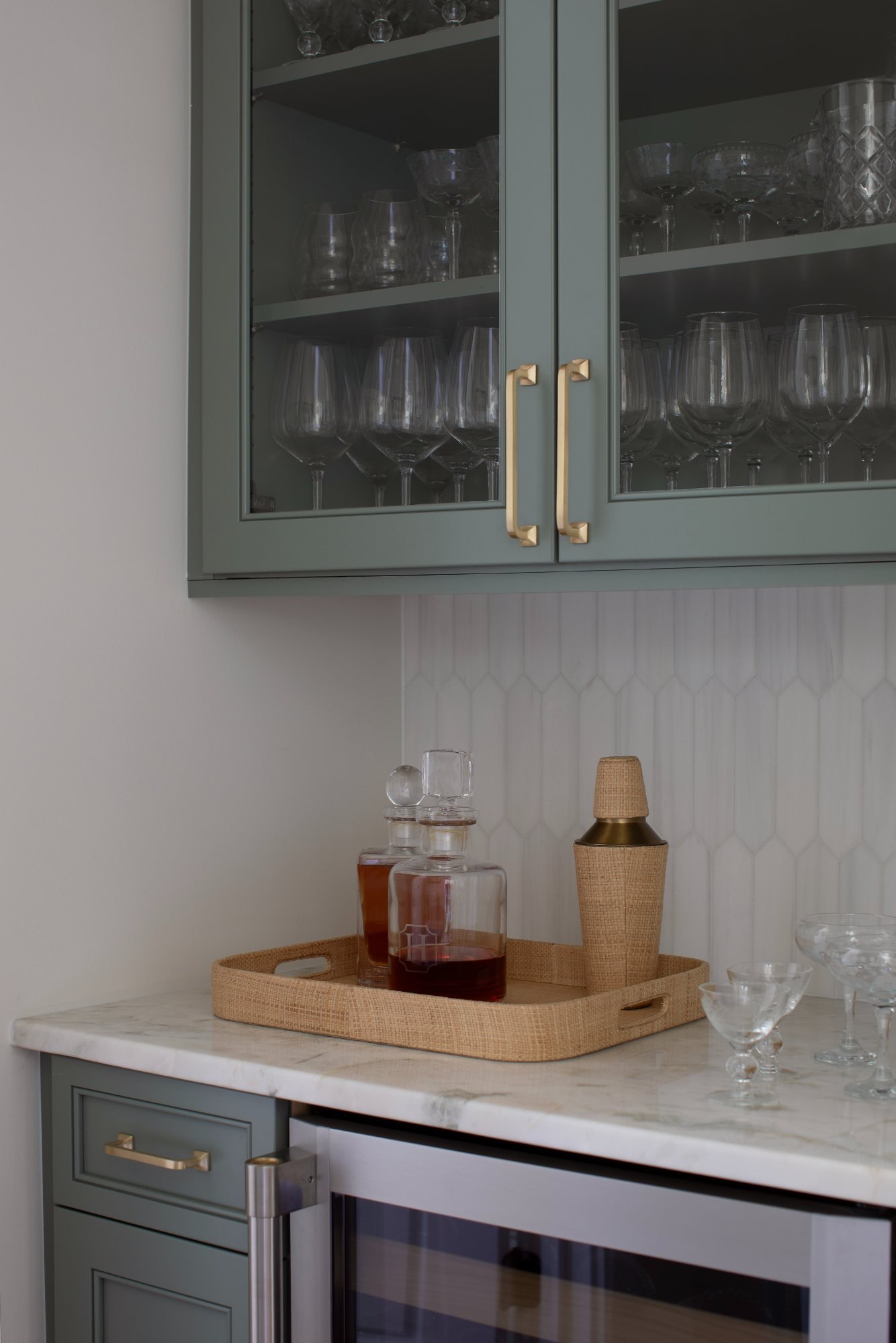 Bar with sage glass-front cabinets, marble counter, and rattan tray of decanters