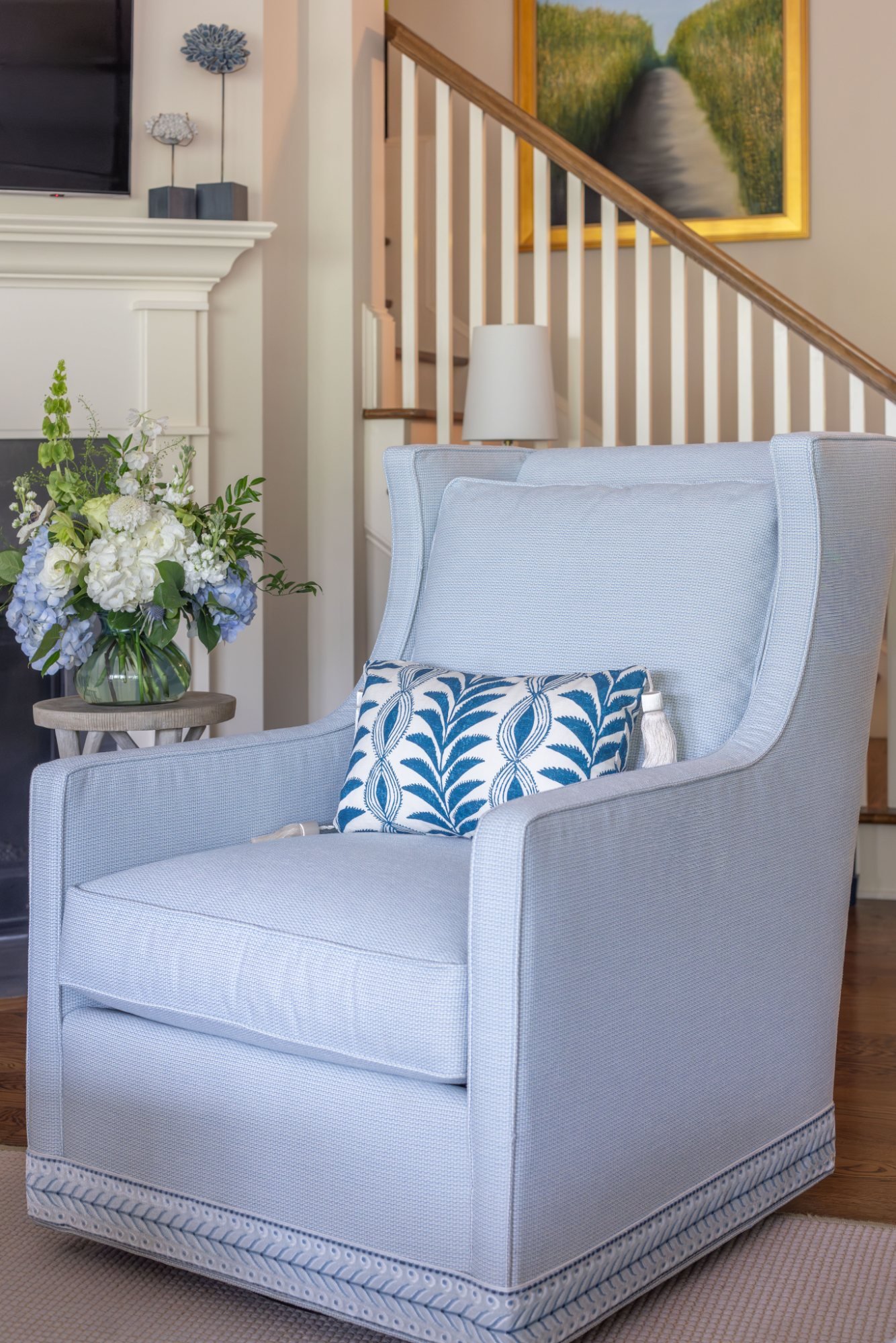 Pale blue wing chair with tassel pillow and braided trim beside the fireplace