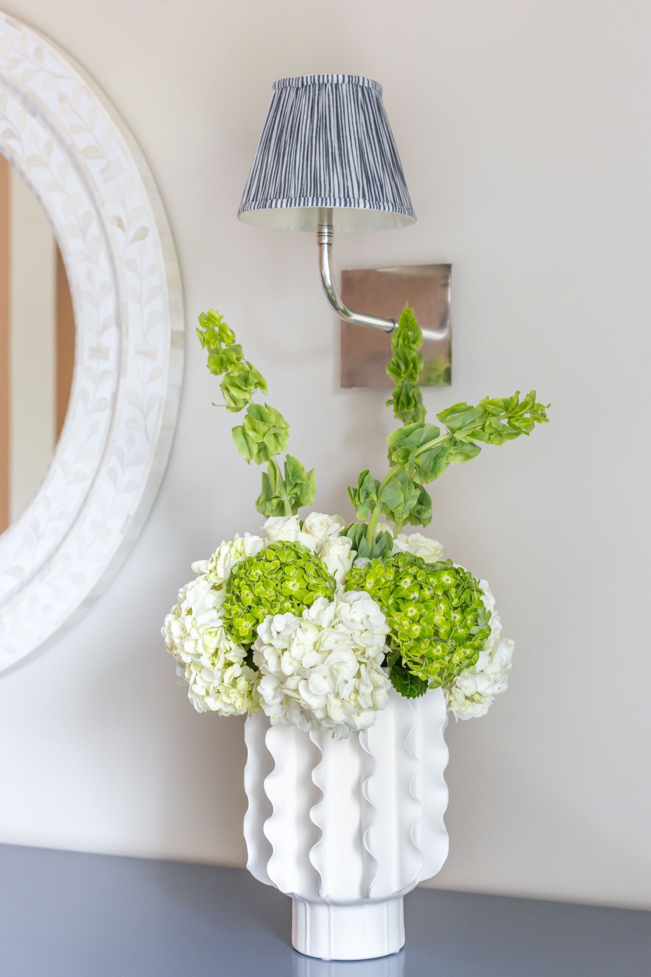 White ruffled vase of hydrangeas and bells of Ireland beneath a pleated sconce