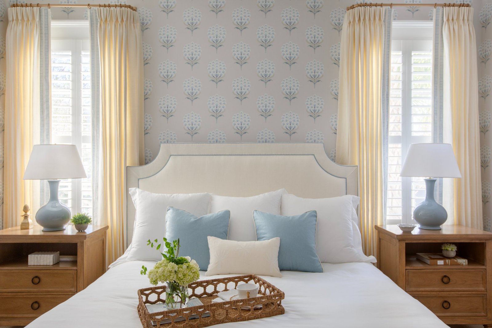 A guest bedroom with blue floral wallpaper, a cream upholstered headboard, pale blue pillows, and a woven breakfast tray of hydrangeas on the bed
