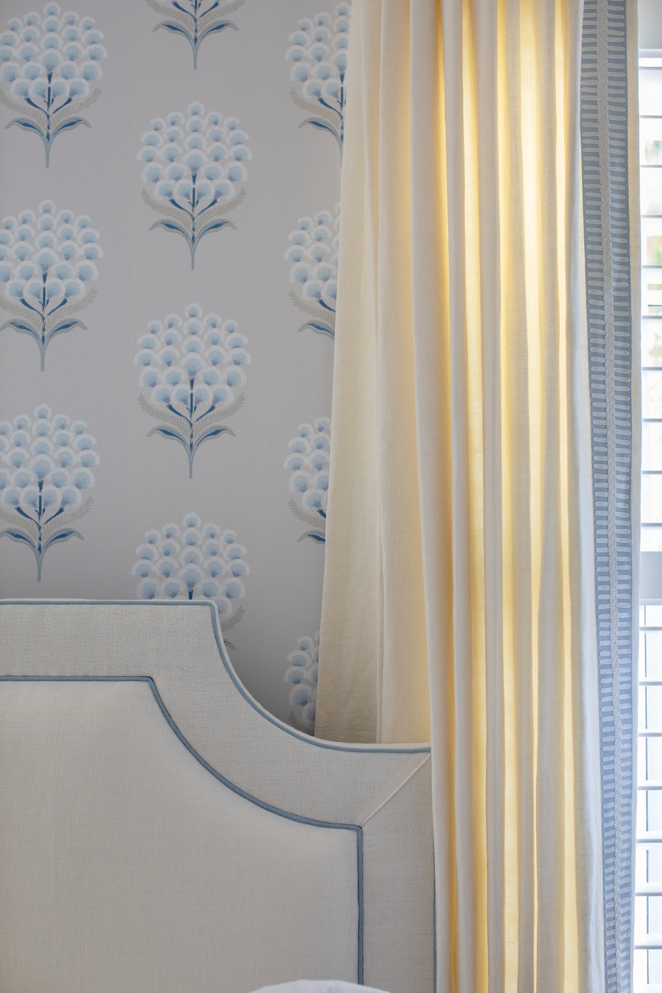 Contrast-piped headboard against block-print paper beside a tape-trimmed linen drape